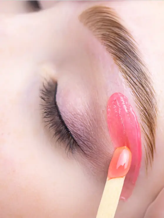 Pink wax is being applied below a woman's eyebrow during an eyebrow waxing treatment
